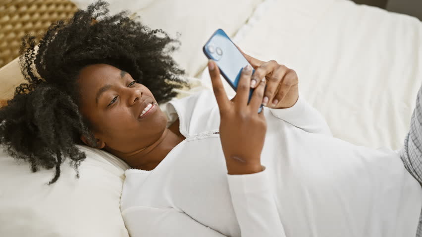 A cheerful african american woman lying in bed using her smartphone in a bedroom setting.