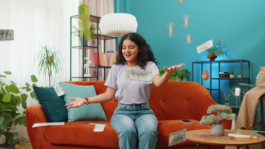 Money cash dollar rain falling on young Indian woman celebrating success, winning, goal achievement, good news lottery jackpot luck. Career grow. Portrait of girl at home apartment living room on sofa