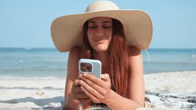 Tanned woman in large hat lies on beach towel by sea, enjoying fun content smartphone. Female looks enthusiastically smartphone screen. Relaxation time lying hot sand tropical island with smartphone. - Powered by Shutterstock - Get 15% off with code: PIKWIZARD15