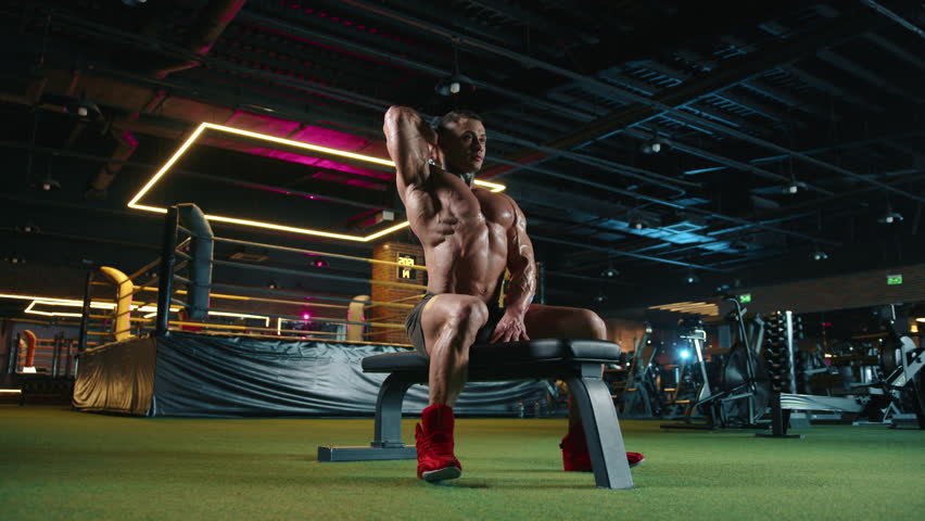 Muscular bodybuilder engages in a focused overhead dumbbell press during an intense workout session at the gym. Fitness, strength, and dedication are highlighted in this powerful moment.