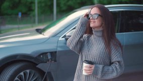 A beautiful young woman in sunglasses, a gray sweater and jeans stands near an electric car while it is charging. The woman holds a cup of coffee in her hands, takes off her glasses and smiles. - Powered by Shutterstock - Get 15% off with code: PIKWIZARD15