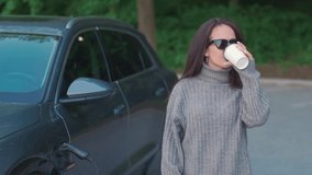 A beautiful young woman in sunglasses, a gray sweater and jeans is standing next to an electric car while it is charging. Woman drinking coffee and smiling. - Powered by Shutterstock - Get 15% off with code: PIKWIZARD15