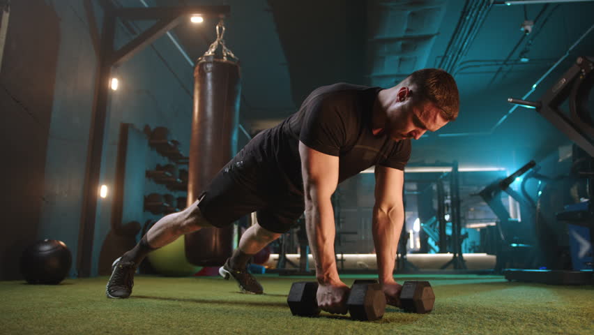 In a wellequipped gym, a man focuses on upper body strength and core stability by performing renegade rows with dumbbells. This workout emphasizes dedication, hard work, and achieving fitness goals