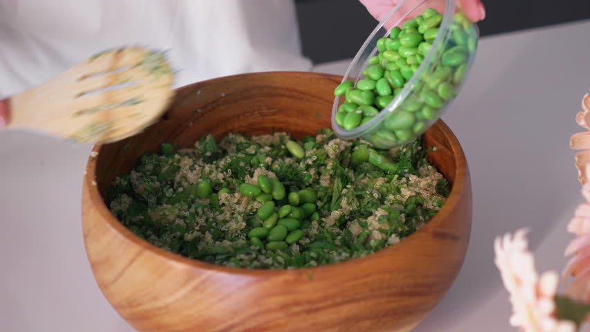 Crop anonymous female hands adding fresh edamame beans to green salad in wooden bowl. Woman is preparing delicious healthy food for lunch in kitchen at home.
