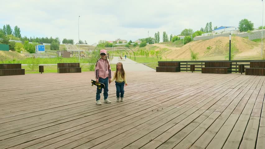 Two little girls, sisters, are walking in the park with a skateboard in their hands. Sports lifestyle. Happy childhood concept