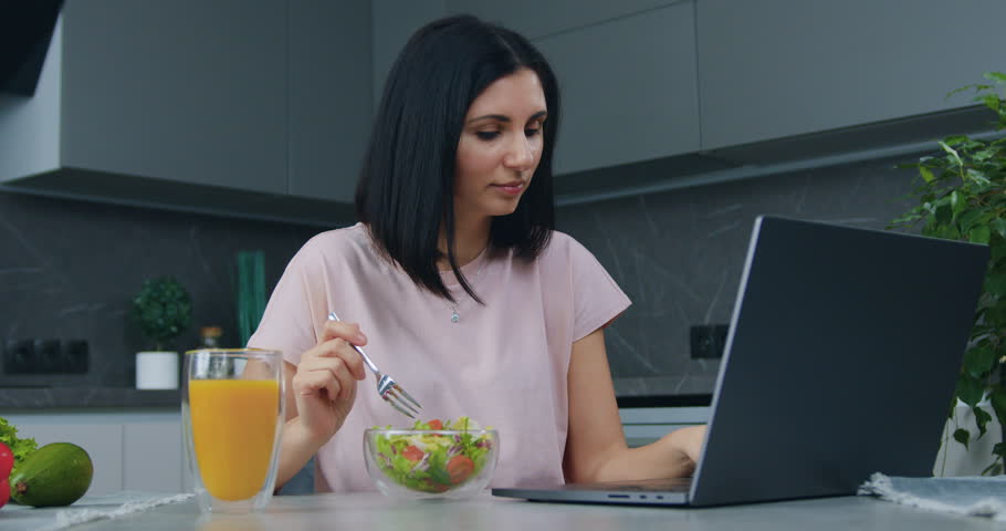 Healthy young woman working on laptop computer and eating vitamin vegetable salad from glass bowl in kitchen