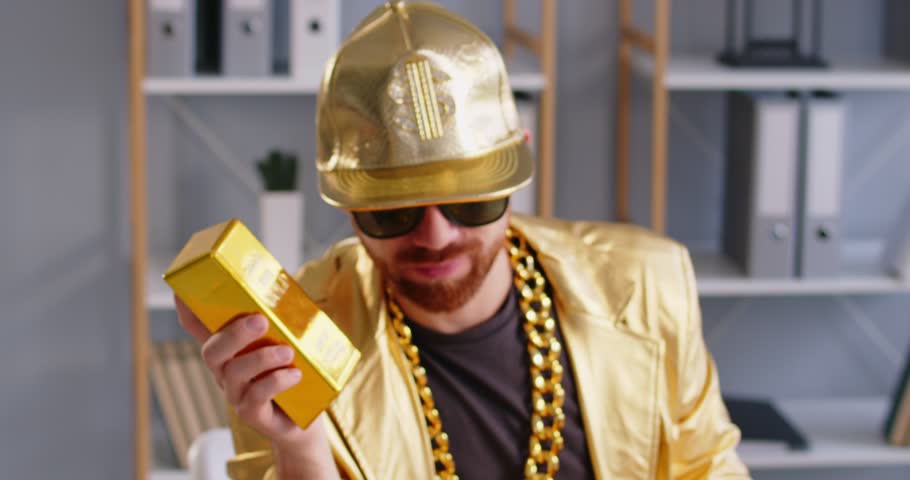 Happy rich man with gold ingot boasting and showing off his wealth. Extravagant eccentric business owner in golden jacket, hat, chain and glasses sitting in office, holding gold bar and smiling