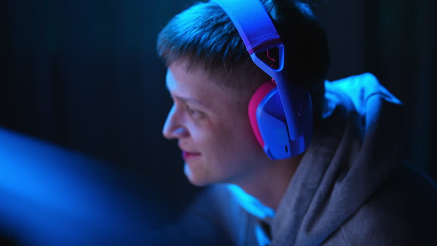 Closeup face of happy excited cyber player wearing gaming headset having fun playing online video games on personal computer in dark room with blue neon light. Concept of modern online entertainment.