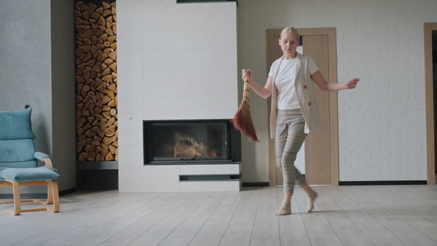 Cheerful middle-aged woman dancing with a broom in her home