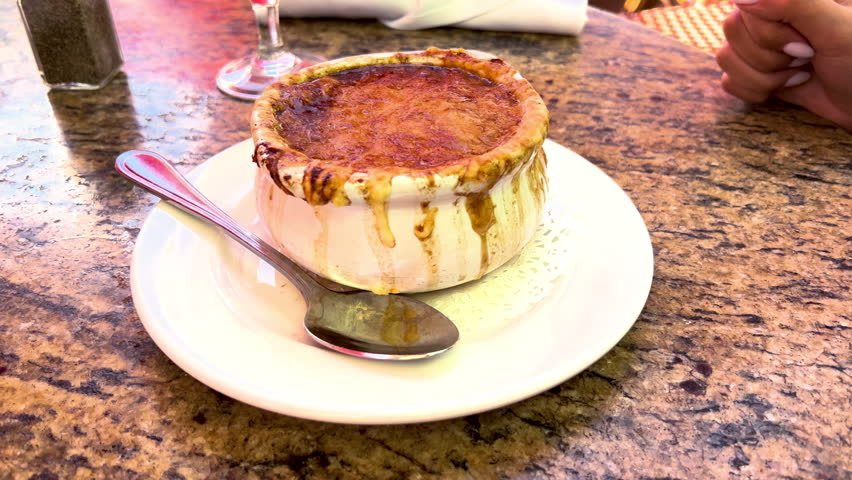 A bowl of boiled-over onion soup with brown stains. Breaking the cheese crust on the top on onion soup with a spoon. A serving of onion soup with a spoon.
