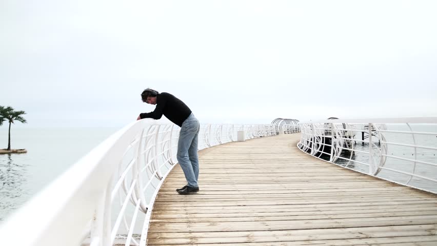 Young guy in a good mood is relaxing outside the city, standing on the pier of a lake, dancing, inspired by the picturesque sea landscape.