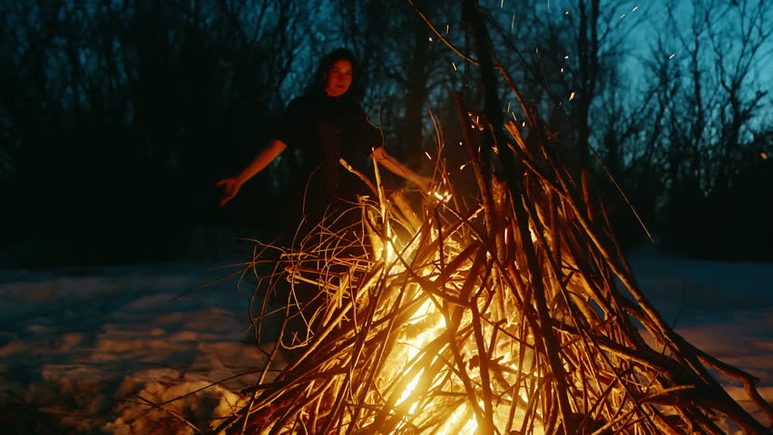 Woman dancing in front of campfire against bare trees. Witch is performing rituals in forest during winter. She is conjuring on Halloween night.
