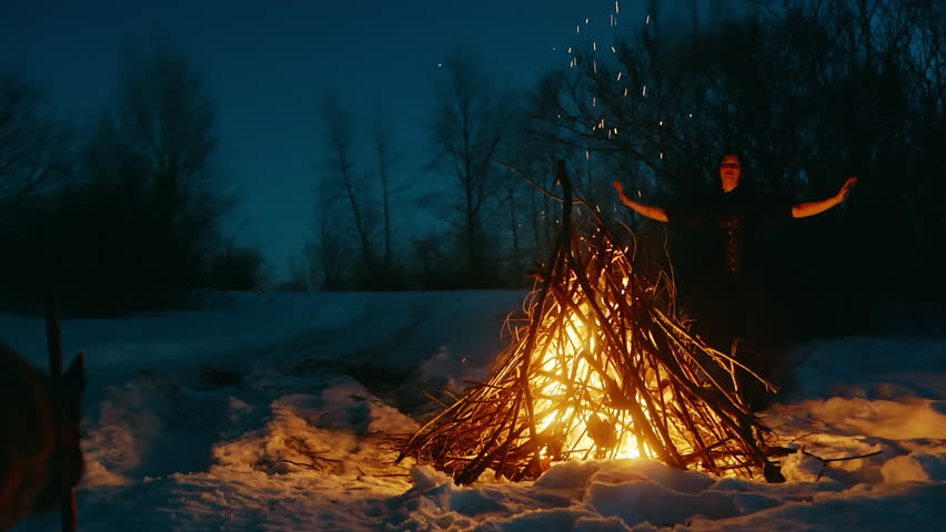 Dog looking at mystical woman dancing in front of bonfire. Witch is performing black magic in forest during winter. They are in dark on snow covered landscape during Halloween night.