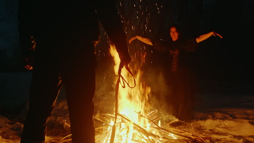 Witch dancing and performing witchcraft at bonfire Man holding pet leash while looking at mysterious magician. They are in dark forest during winter on Halloween night.