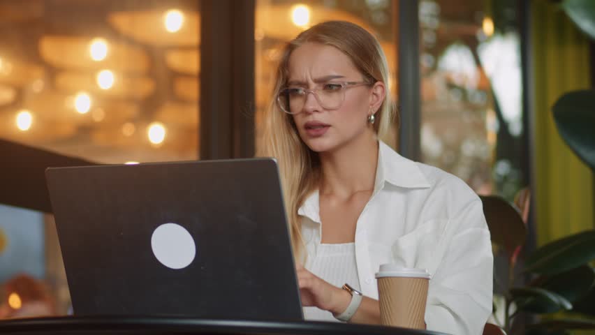 Beautifully decorated cafe vibrant greenery blonde business woman in glasses working on laptop cup of coffee nearby. Caucasian businesswoman wearing glasses sits in chic cafe adorned with lush plants.