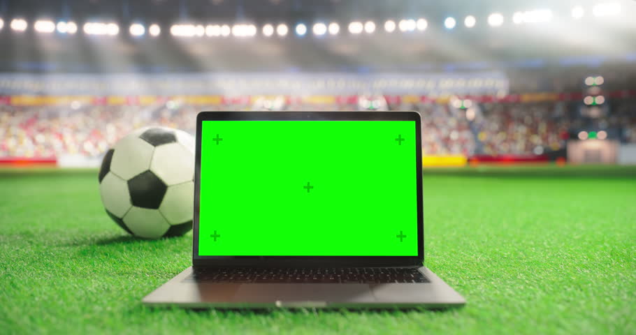 Laptop Computer with a Green Screen Placed on Grass Next to a Soccer Ball in a Stadium, Suitable for Mockups and Sports Marketing. Ideal Setup for Showcasing Digital Content in a Sports Environment