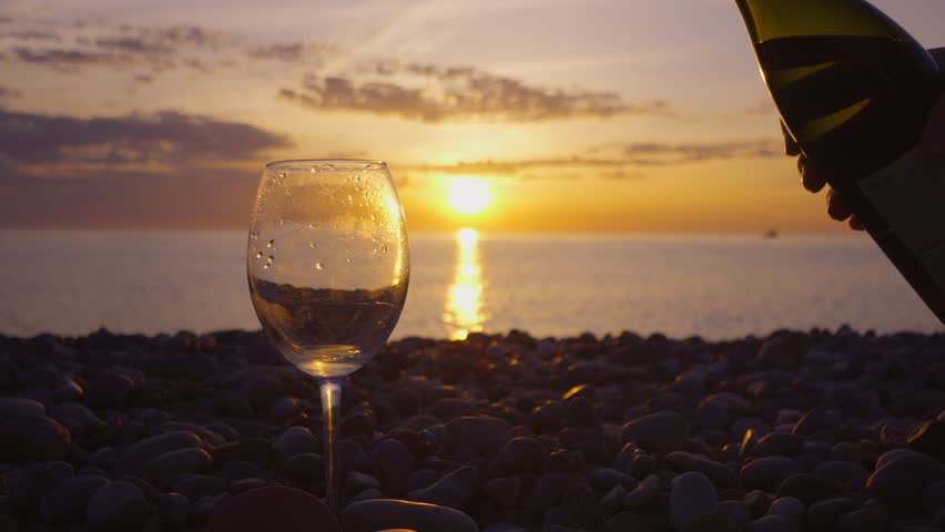 Slow Motion white wine poured into glass from bottle on rocky seashore at sunset orange sun. Pouring glass of white wine against backdrop of sea and sun