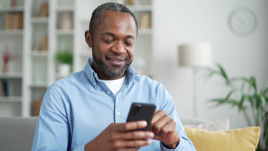 Mature african american man using smartphone sitting on sofa in living room at home. Smiling male browses social networks, does shopping online, chats with a friend, writes or reads messages. Close up - Powered by Shutterstock - Get 15% off with code: PIKWIZARD15