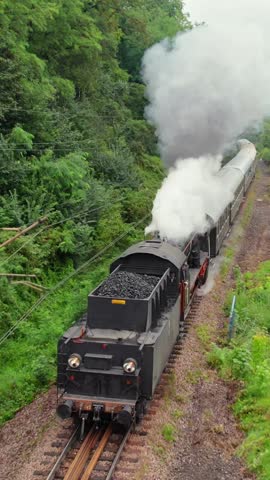 Aerial drone view of the moving steam train in countryside. Vintage coal steam locomotive with smoke on trail. Retro transport and travel. Historic train on railroad on a full speed.