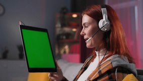 video call, young woman with a headset uses modern technology to communicate with friends using a camera on an electronic gadget while sitting on the sofa in the room in the evening, green screen