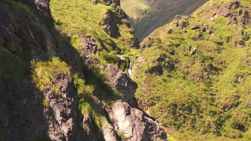 Drone image emerging from behind rocks and revealing a beautiful waterfall nestled in the Andes mountains.