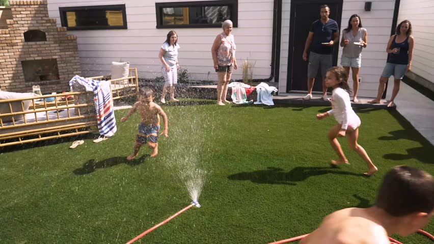 Playful kids running through sprinkler in sunny summer backyard