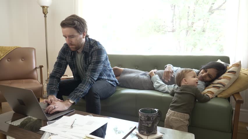 Man working at laptop on sofa by resting pregnant wife and toddler son