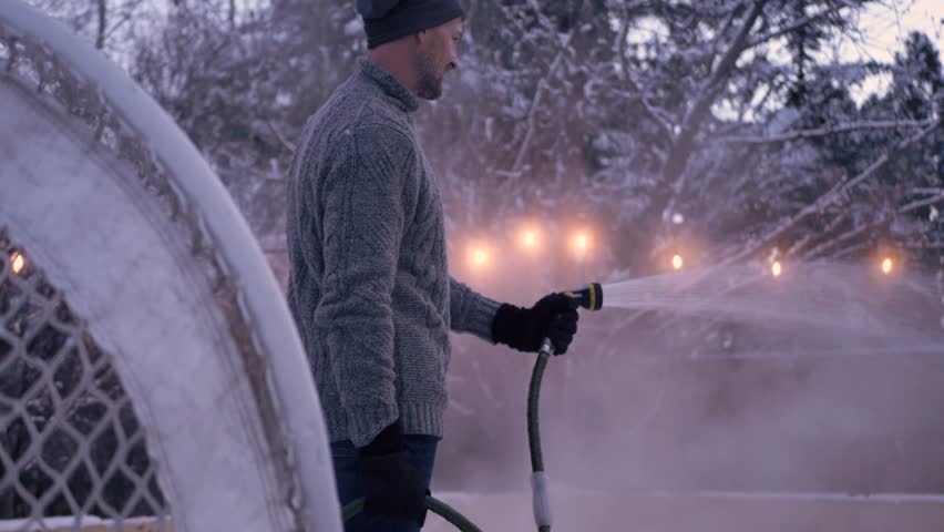 Man with hose spraying ice hockey rink in snowy winter backyard