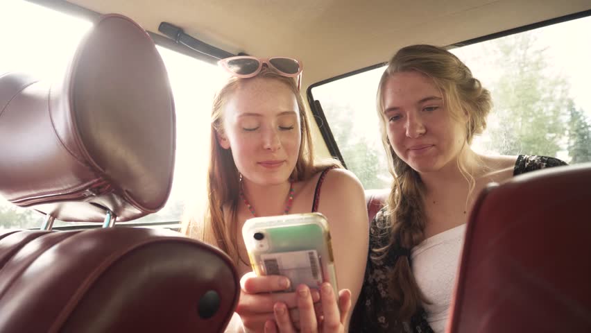 Happy teenage girl friends using smart phone in back seat of sunny car