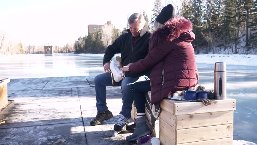 Senior man helping wife tie ice skate shoelaces at frozen park pond