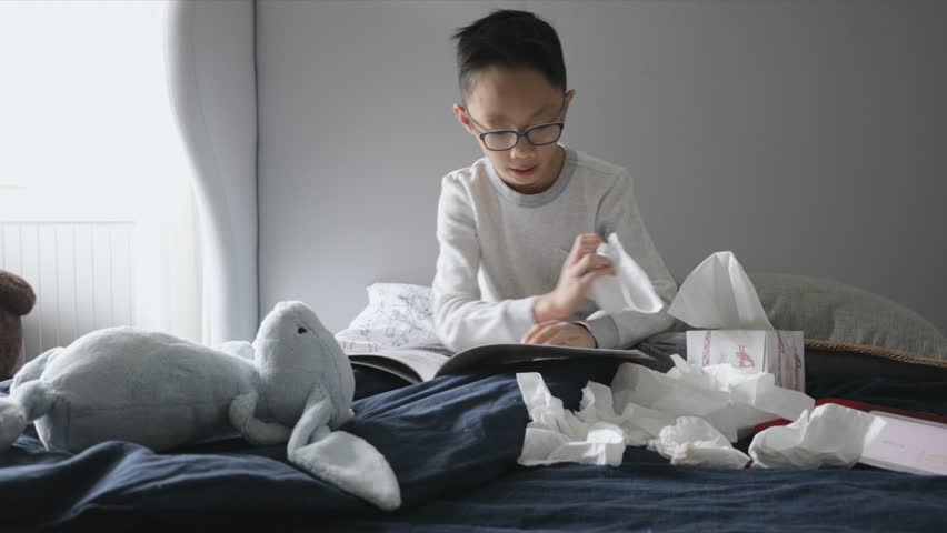 Boy with cold reading book and blowing nose with tissue on bed