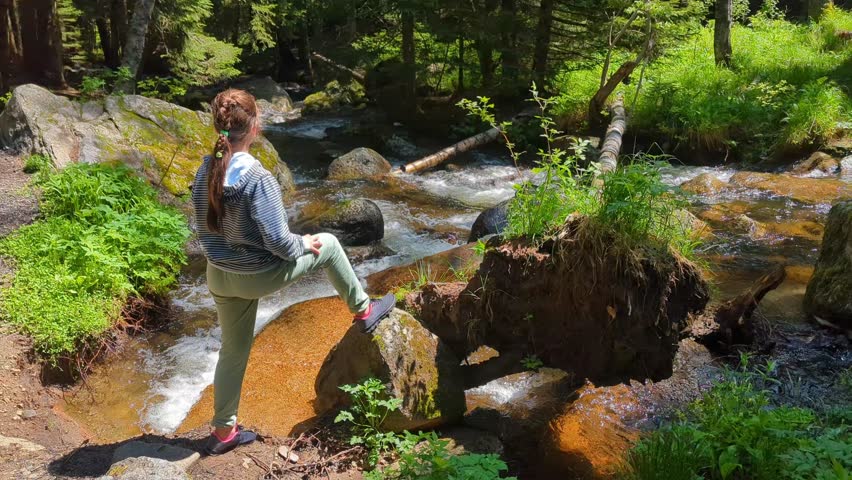A girl stands on rocks near a mountain river with a fallen tree creating a natural bridge, surrounded by the forest. She admires the wilderness and rests after a hike.
