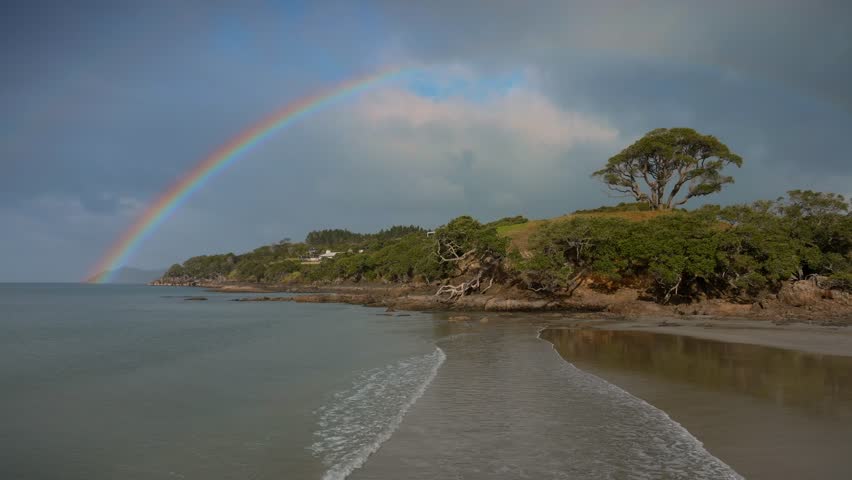 Aerial: Calm ocean waves and Rainbow over sandy coastline and pohutukawa tree on a hill in Bream Bay, Waipu, Northland, New Zealand.