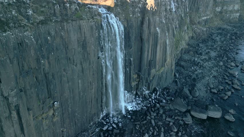 Waterfall half speed slow motion in shadow falling down sheer cliff to frosted rocky beach below in winter at Kilt Rock Waterfall, Isle of Skye, Western Highlands, Scotland, UK