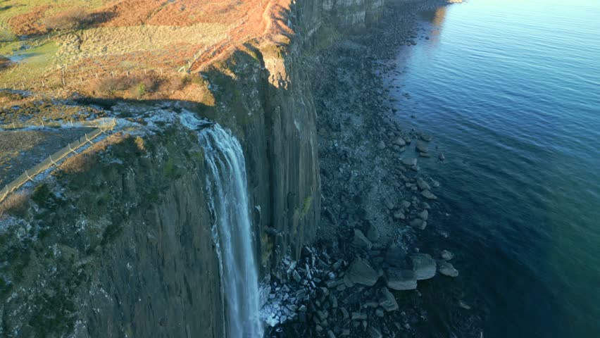 Waterfall in shadow cascading down steep cliff to frosty and stony beach, early morning in winter with sunlit moorland top at Kilt Rock Waterfall, Isle of Skye, Western Highlands, Scotland, UK