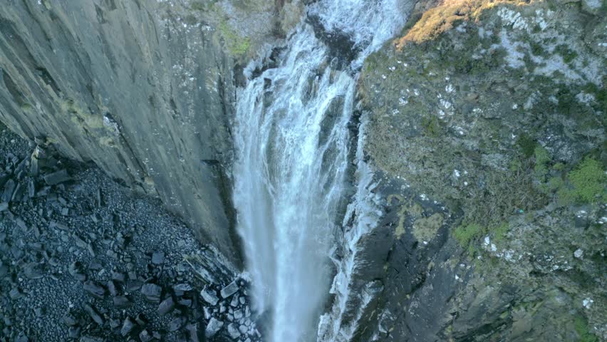 Top down view of waterfall cascading over icy steep cliff to frosted dark stony beach below. Early morning in winter slow orbit at Kilt Rock Waterfall, Isle of Skye, Western Highlands, Scotland, UK