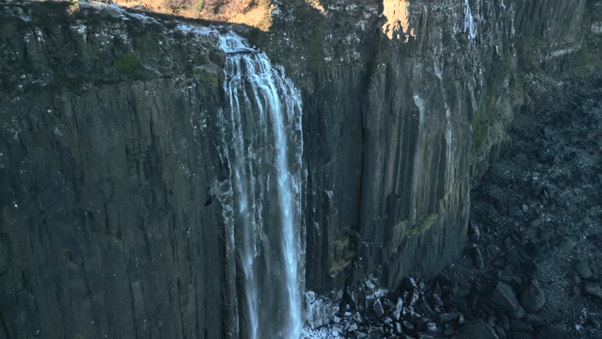 Waterfall half speed slow motion orbit in shadow falling down sheer cliff to frosted rocky beach below in winter at Kilt Rock Waterfall, Isle of Skye, Western Highlands, Scotland, UK