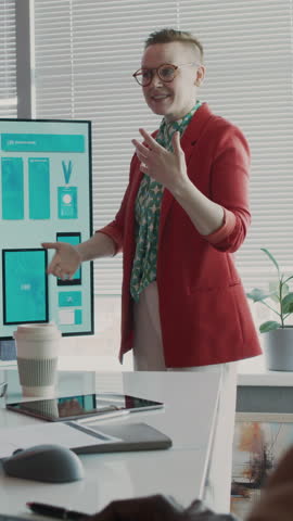 Medium vertical shot of charismatic Caucasian female brand manager in red jacket presenting new merchandise design for marketing campaign on multimedia screen at corporate meeting