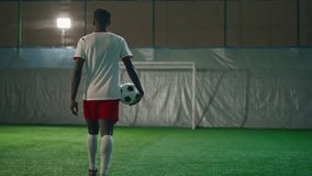 Young African American soccer player walking on artificial field indoors, holding football ball in hand, back view. Professional sport and sportive hobby, black guy training in soccer club, forward - Powered by Shutterstock - Get 15% off with code: PIKWIZARD15