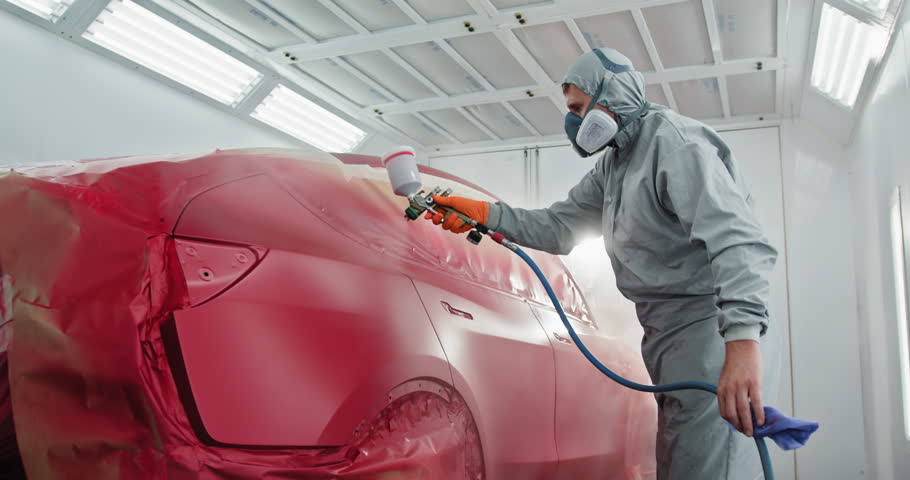 A car painter in a protective suit and mask is spray painting a car in a specialized booth in an auto body shop. Auto Painter Applying Paint in Spray Booth. Concept of automotive and car body repair