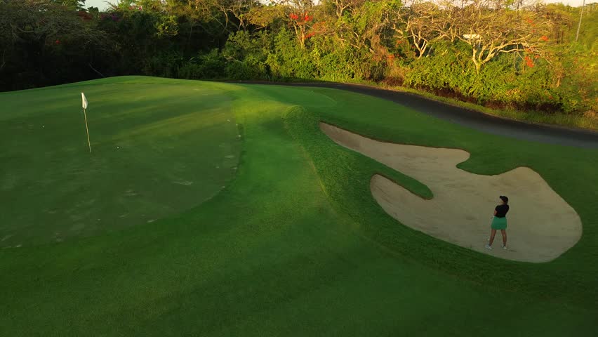 An aerial view of a young woman playing professional golf as she hits a golf club out of the sand from down to up. The ball flies up on the hill, the woman jumps up to see if she hits the hole.