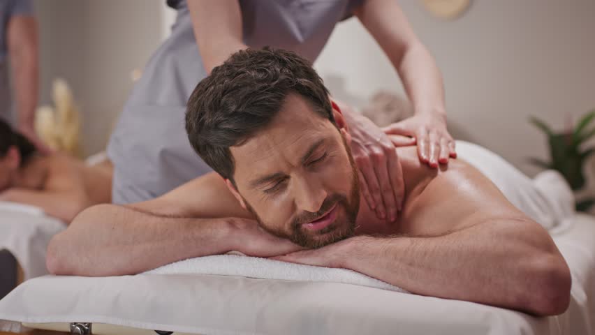Middle-aged man receiving massage in spa salon. Resting while female massauer kneading his shoulder. Proffesional therapist taking care about patients body. Providing rejuvenation for skin. Treatment.