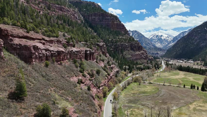 Aerial View of Highway Winding Through the Mountains, rocky jagged cliffs on one side of the road with pine trees, partial blue sky with clouds in background, Ouray Colorado