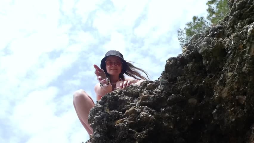 Girl in pink swimsuit and glasses on a rock near the water gives a hand while on top