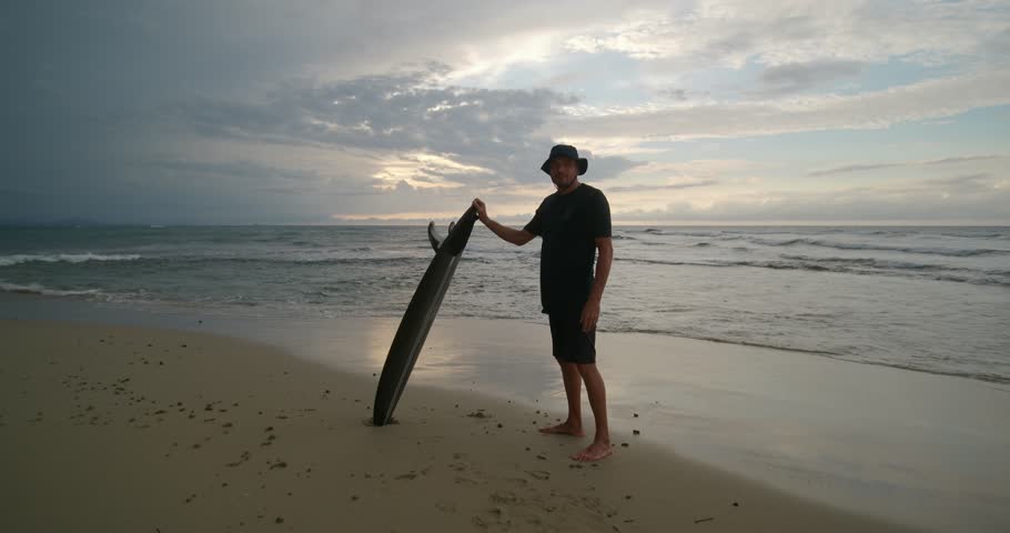 Surfer with surfboard stands on ocean beach observing waves at sunset. 