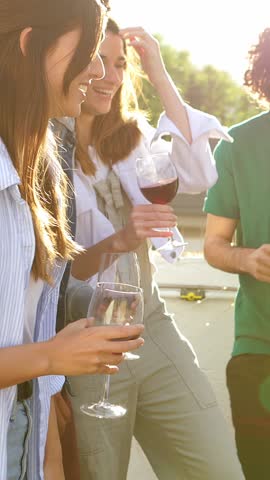 Young diverse group of friends enjoying summer party at rooftop terrace, dancing and drinking red wine. Happiness and celebration concept.