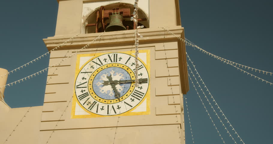 A large clock with Roman numerals and a bell on top is attached to a building, decorated with strings of lights under a clear, blue sky.