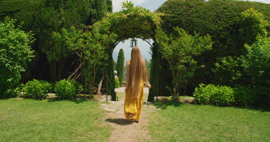 A woman walks through a lush archway garden in summer sunny day. Her long blonde hair cascades behind her in the sunlight.