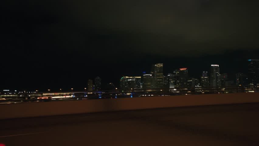Point of view of car driving plate on Miami highway illuminated at night. POV car windshield view of Miami Skyline modern buildings illuminated at night