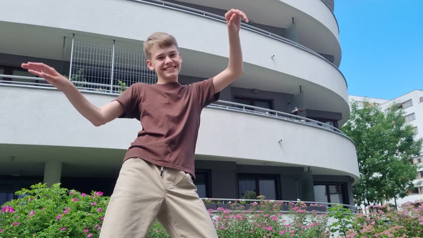 Teenager dancing energetically outdoors in residential area, buildings and greenery in background.
